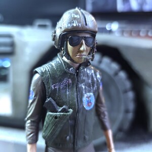 May include: A close-up of a toy action figure wearing a brown helmet with a faded inscription, a brown leather jacket with a patch that says "FERRO", and a blue patch with a symbol on the left side of the chest. The figure is holding a gun in a holster on the right side of the jacket.