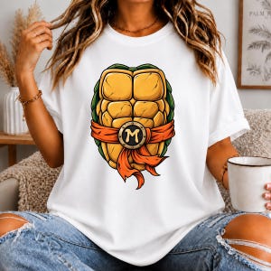 Op de afbeelding: Een wit T-shirt met korte mouwen en een grafische print van een geel schildpadschild met groene randen, vastgebonden met een oranje riem met een ronde gesp in het midden waarop de letter M in een vet, gestileerd lettertype staat.