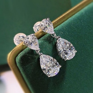 May include: A pair of elegant drop earrings featuring sparkling, pear-shaped cubic zirconia stones. The earrings have a silver-colored setting and are displayed against a green velvet background. The earrings are designed to dangle gracefully.