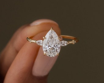 May include: A gold ring featuring a pear-shaped diamond center stone. The ring has a delicate band with smaller accent stones and a classic, elegant design. The ring is shown against a neutral background.