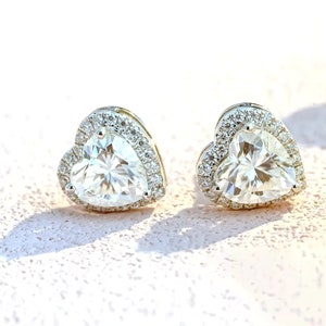 May include: A pair of heart-shaped stud earrings. Each earring features a large, clear, heart-cut gemstone surrounded by a halo of smaller, sparkling stones. The earrings are set in a silver-colored metal and are displayed on a light pink surface.
