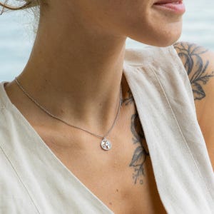 Puede incluir: Un collar de cadena de plata con un pequeño colgante redondo. El colgante tiene una superficie texturizada y parece estar hecho de metal.