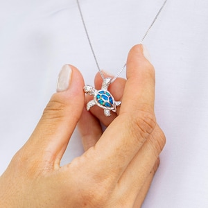 Puede incluir: Un collar plateado con un colgante de tortuga. El colgante tiene un caparazón azul y blanco similar al ópalo y detalles en las aletas y la cabeza. El collar lo sostiene una persona. La cadena es delicada y plateada.