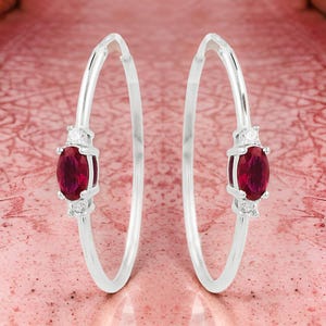 May include: A pair of silver hoop earrings with a red oval gemstone and two smaller white gemstones on each earring.