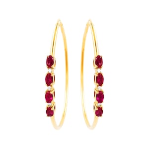 May include: A pair of gold hoop earrings, each adorned with five oval-cut, deep red gemstones and small, sparkling accent stones. The earrings are set against a white background, highlighting their elegant design and vibrant color.