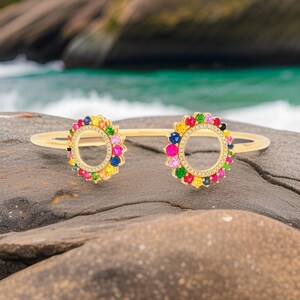 May include: A gold bangle bracelet with two circular designs. Each circle is adorned with a halo of colorful gemstones, including pink, red, yellow, green, blue, and purple.