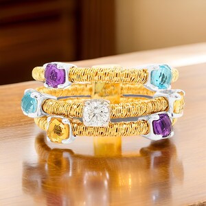 May include: A gold ring with a woven band and five gemstones. The gemstones are a round diamond, a square amethyst, a square topaz, a square citrine, and a square aquamarine.