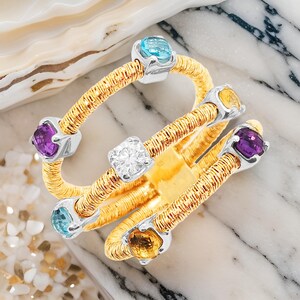 A gold ring with multiple bands set with a variety of gemstones including a large clear diamond, a smaller blue gemstone, a smaller yellow gemstone, and two smaller purple gemstones. Italian design multi gemstone ring.