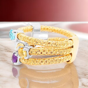 May include: A gold ring with three bands, each featuring a different coloured gemstone. The gemstones are a light blue topaz, a yellow citrine, and a purple amethyst.