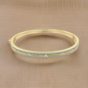 May include: A gold-colored bangle bracelet with a textured center band and a row of small, clear stones. The bracelet has a hinged clasp and is set against a neutral background.