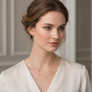 Puede incluir: Una mujer lleva un delicado collar de oro con m&uacute;ltiples piedras preciosas blancas, peque&ntilde;as y redondas. El collar destaca sobre una blusa blanca. La mujer tiene la piel clara y cabello casta&ntilde;o.
