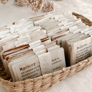 May include: A wicker basket filled with small, folded pieces of paper, each with a different word or phrase written on it. The words include "Mystery", "Believe", and "What is hidden".