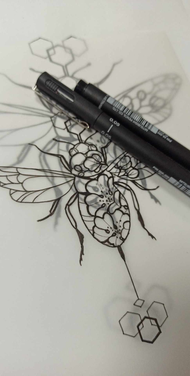 May include: A black and white line drawing of a bee with geometric shapes surrounding it. Two black pens are lying on top of the drawing. The pens are labeled "0.05" and "0.3", and "MITSUBISHI PENCIL CO. LTD."