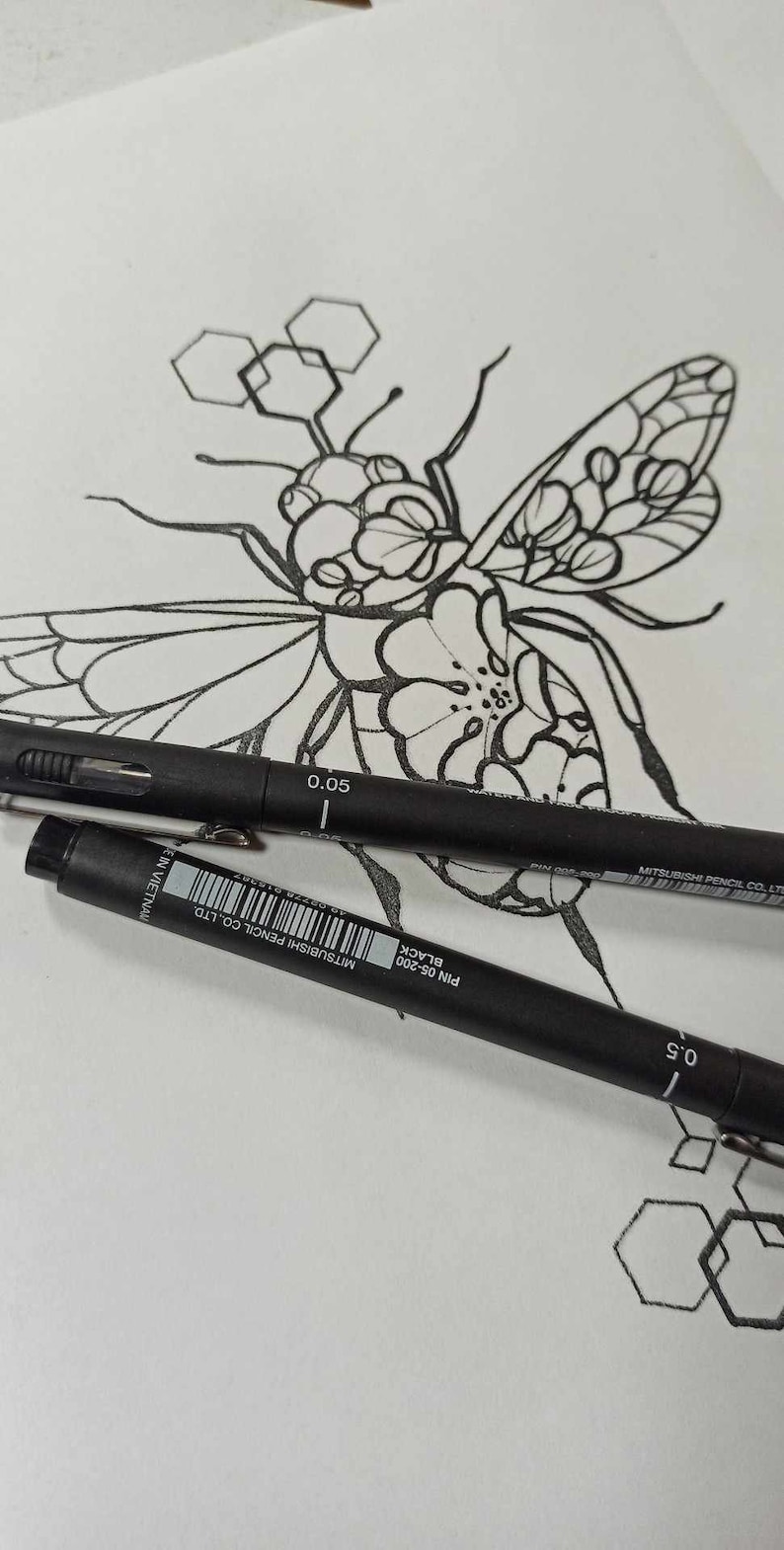 May include: A black and white line drawing of a bee with geometric patterns on its wings and body. The bee is drawn on a white sheet of paper with two black pens lying on top of the drawing. The pens are labeled "Pin 0.5-200 Black" and "Mitsubishi Pencil Co. Ltd."
