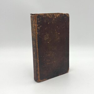 May include: An antique hardcover book with a weathered, dark brown leather cover. The binding shows significant signs of wear, including frayed edges, scuffs, and discoloration on the spine and corners, indicating age and extensive handling.