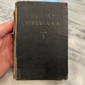 Può includere: Un libro vintage con copertina rigida nera intitolato TODAY WITH GOD in lettere dorate sulla copertina anteriore, con un piccolo simbolo di croce dorata sotto il testo. Il libro presenta dorso e bordi usurati e sfilacciati dal tempo e dall'uso.