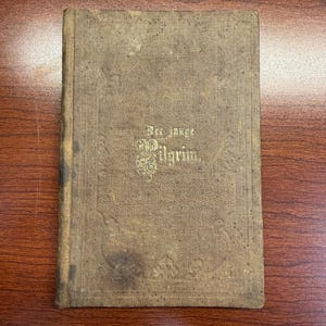 Könnte beinhalten: Ein antikes Buch mit festem Einband aus verwittertem, beigefarbenem Stoff. Der Vorderdeckel zeigt geprägte dekorative Ränder und den goldgeprägten Schriftzug "Der junge Pilgrim" in der Mitte, der deutliche Alters- und Gebrauchsspuren aufweist.