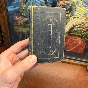 Pode incluir: Um pequeno livro antigo retangular com uma capa preta desgastada, apresentando bordas decorativas douradas em relevo e um painel central retangular. As bordas do livro mostram sinais de idade e desgaste, com desfiamento visível na lombada.