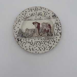 May include: A round ceramic plate with a cream-coloured base featuring a black alphabet border separated by small flower icons. The centre displays a historical illustration titled BIBLE PICTURES and THE DESTRUCTION OF PHARAOH with figures.