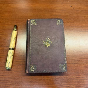 Op de afbeelding: Een klein, rechthoekig, donkerpaars hardcover boek met gouden bloemmotieven in de hoeken en een centraal gouden bloemembleem, naast een lichte, cilindrische houten pen met donkere, onregelmatige nerven en gouden metalen accenten.