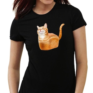 May include: Black t-shirt featuring a whimsical graphic of a cat shaped like a loaf of bread. The cat is orange and white, with a smiling expression. The shirt is made of a soft material and has short sleeves.