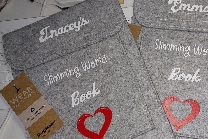 May include: Two gray felt bags with a white heart and the text "Slimming World Book" on the front. The bags have a brown tag that says "Wear Don't Waste" and "BagBase".
