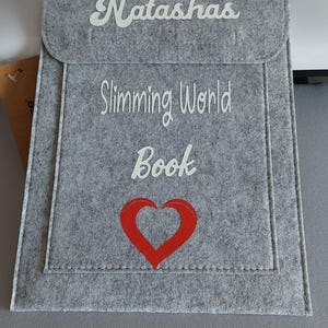 White htv vinyl with large name at the front flap top of the folder.
then the word slimming world book and a large red hollow heart on the bottom part of the folder.
grey stitching and soft felt feel. 
large main compartment & small pocket at front.
