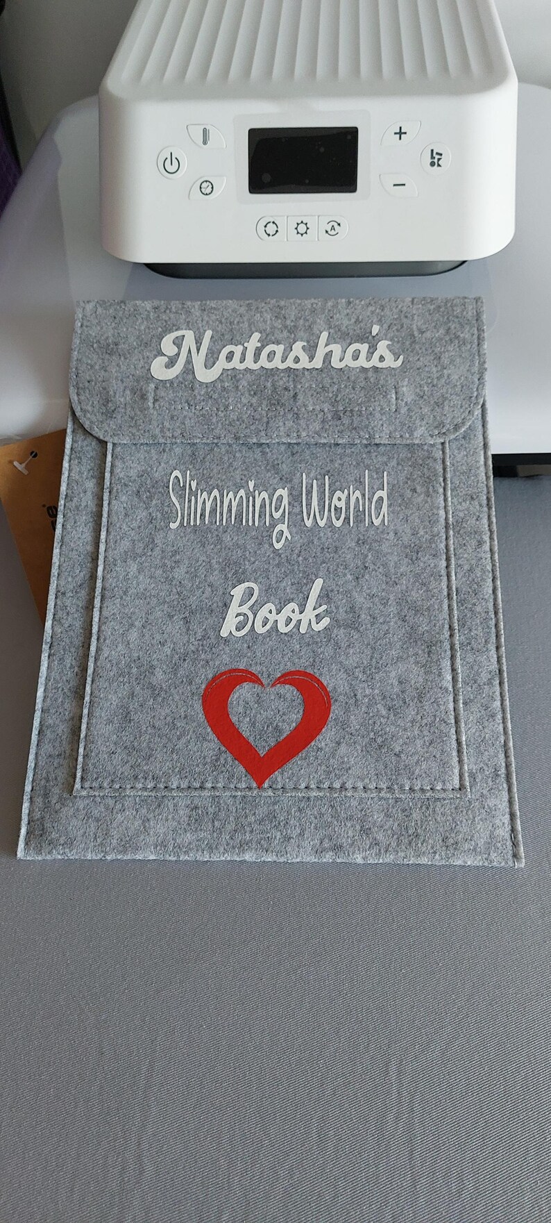 White htv vinyl with large name at the front flap top of the folder.
then the word slimming world book and a large red hollow heart on the bottom part of the folder.
grey stitching and soft felt feel. 
large main compartment & small pocket at front.