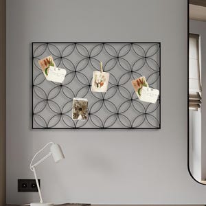 Peut inclure: Un tableau mémo en métal noir avec un motif géométrique circulaire, contenant plusieurs petites cartes. Le tableau est fixé sur un mur gris clair au-dessus d'un bureau en bois, avec une lampe blanche et une chaise. La pièce a un design moderne et minimaliste.