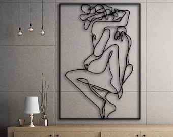 May include: A black metal wall art sculpture depicting two figures in an abstract, intertwined pose. The sculpture is framed in a black square frame and hangs on a light gray wall above a wooden cabinet.