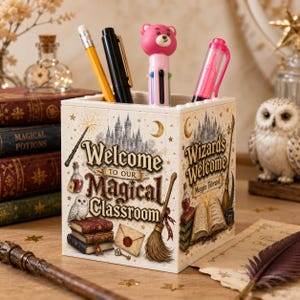 May include: A square pencil holder constructed from building blocks, featuring printed graphics of a castle, a broomstick, books, and an owl. The text reads Welcome to our Magical Classroom and Wizards Welcome Muggles Tolerated. It holds four pens.
