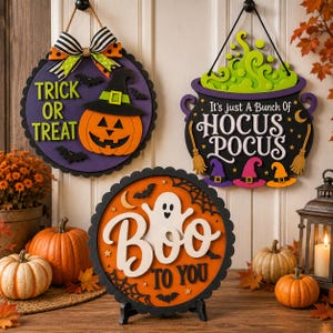 May include: Three Halloween-themed wall signs. One purple circular sign features a pumpkin, bats, and the text "TRICK OR TREAT". A black cauldron sign displays green bubbling liquid and the text "It's just A Bunch Of HOCUS POCUS". An orange circular sign shows a ghost, spiderwebs, and "Boo TO YOU".