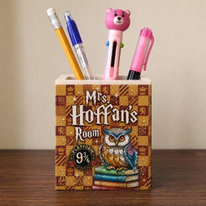 May include: A rectangular pencil holder featuring a gold and brown checkered pattern with the text Mrs. Hoffan's Room and Platform 9 3/4. The design includes an owl perched on a stack of books. Four pens and pencils are stored inside the container.