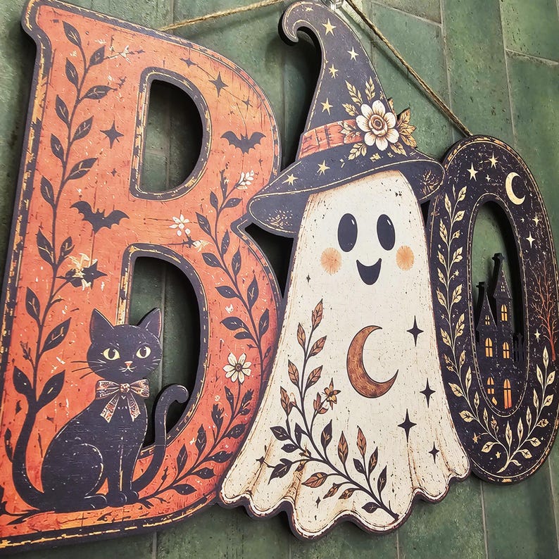 May include: A decorative wall sign spelling BOO with a black cat on the letter B, a ghost wearing a witch hat in the center, and a haunted house silhouette inside the letter O. The design features orange and black colors with floral motifs.