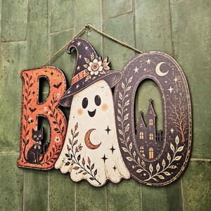May include: A decorative wall hanging spelling the word BOO. The letter B is orange with a black cat illustration. The letter O is a white ghost wearing a witch hat with a crescent moon. The final O is black with a haunted house silhouette.