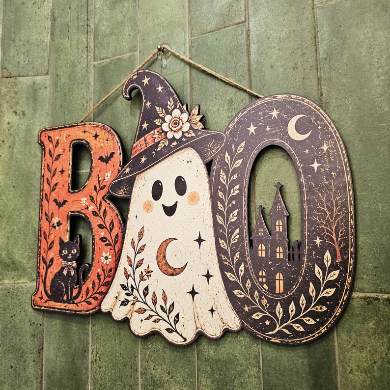 May include: A decorative wall hanging spelling the word BOO. The letter B is orange with a black cat illustration. The letter O is a white ghost wearing a witch hat with a crescent moon. The final O is black with a haunted house silhouette.