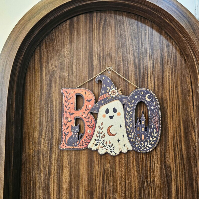 May include: A decorative hanging sign spelling the word BOO. The letter B features a black cat and bats on an orange background. The letter O is replaced by a cream-colored ghost wearing a witch hat. The final O depicts a dark haunted house.