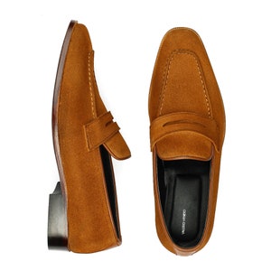 May include: A pair of brown suede loafers with a leather sole. The loafers have a single strap across the top and the brand name "VALERO VINELLO" is printed on the insole.