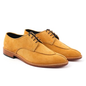 May include: Mustard-coloured suede dress shoes with brown laces. The shoes have a classic design with a stitched sole and a low heel. The shoes are displayed on a white background.