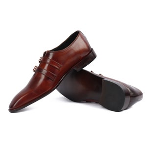 May include: A pair of brown leather monk strap shoes with two buckles on each shoe. The shoes are on a white background.