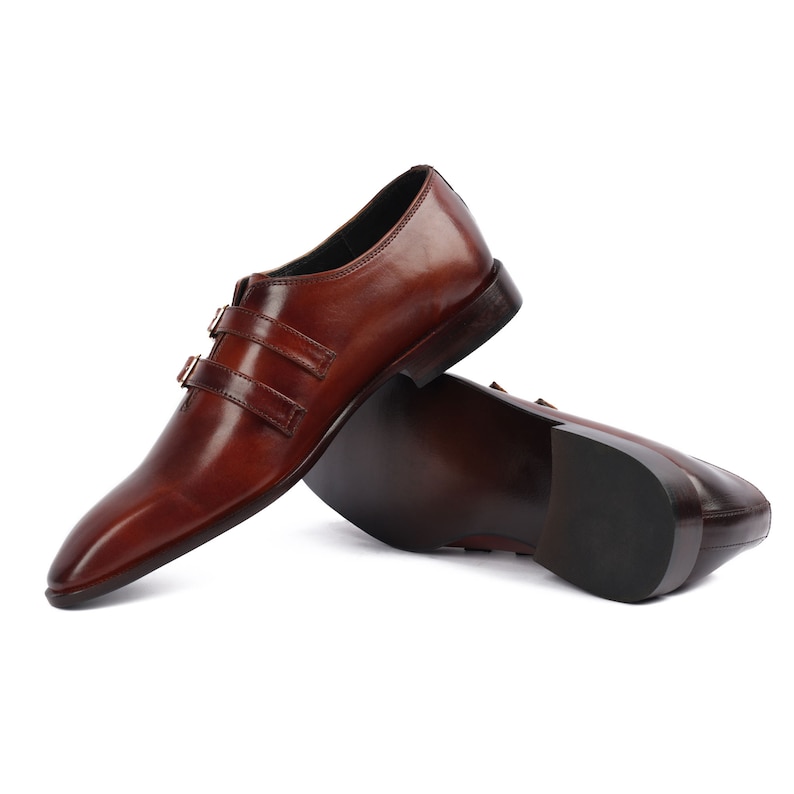 May include: A pair of brown leather monk strap shoes with two buckles on each shoe. The shoes are on a white background.