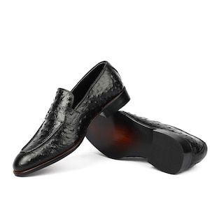 May include: A pair of black leather loafers with an ostrich print. The loafers are stacked on top of each other, with the top loafer's sole facing the camera.