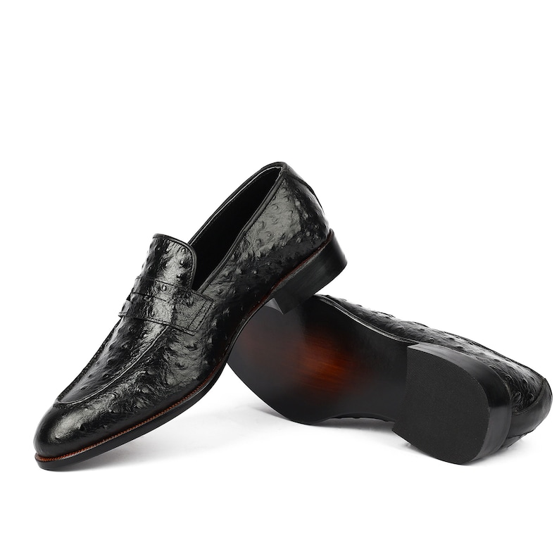May include: A pair of black leather loafers with an ostrich print. The loafers are stacked on top of each other, with the top loafer's sole facing the camera.