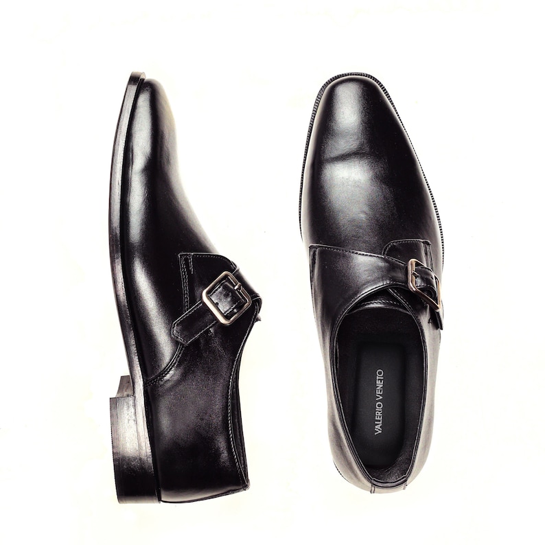 May include: A pair of black leather monk strap shoes with a silver buckle. The shoes have a pointed toe and a low heel. The brand name "VALERIO VENETO" is printed on the inside of the right shoe.