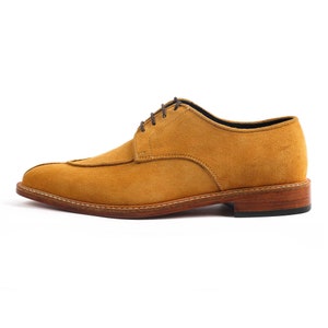 May include: A single, ochre-coloured suede dress shoe with a stitched design and brown laces. The shoe has a leather sole and a small heel. The shoe is designed for formal occasions.