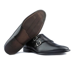 May include: Black leather dress shoes with double monk strap buckles. The sole is brown with a textured pattern. The shoes are displayed on a white background.