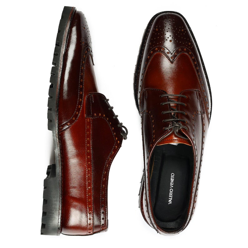 Pu&ograve; includere: Un paio di scarpe da uomo in pelle marrone con punta a brogue e chiusura con lacci. Le scarpe hanno un design a punta alare e un motivo traforato. Il nome del marchio "VALERIO VENETO" &egrave; visibile all'interno di una scarpa.