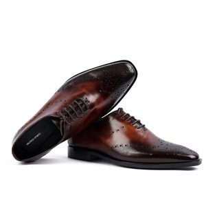 May include: A pair of brown leather lace-up shoes with a brogue design and a pointed toe. The shoes have a dark brown leather upper and a black leather sole. The shoes are made by the brand "VALERO VIANTO".