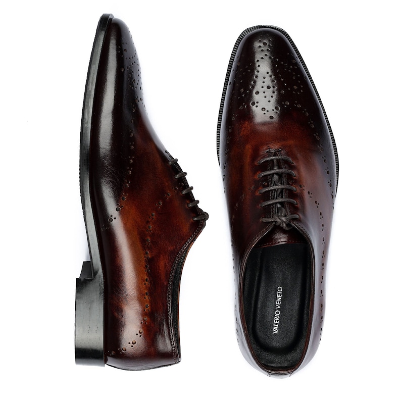 May include: A pair of brown leather lace-up shoes with a perforated design. The shoes have a black leather sole and a black leather lining. The brand name "VALERIO VENETO" is printed on the inside of the right shoe.