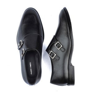 May include: A pair of black leather double monk strap shoes. The shoes have silver-colored buckles and a low heel. The brand name "VALERIO VENETO" is visible on the insole of one shoe. The shoes are displayed on a white background.